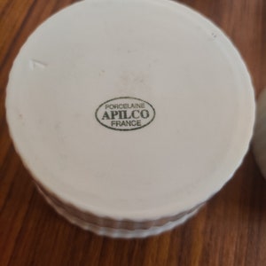 May include: White porcelain ramekin with a ridged exterior. The bottom of the ramekin features a green oval stamp with the text "PORCELAINE APILCO FRANCE". The ramekin is designed for baking and serving individual portions.