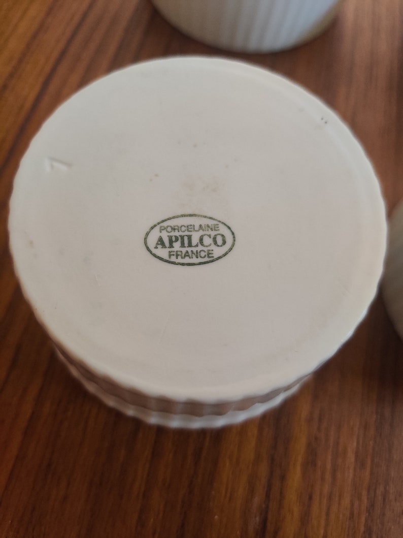 May include: White porcelain ramekin with a ridged exterior. The bottom of the ramekin features a green oval stamp with the text "PORCELAINE APILCO FRANCE". The ramekin is designed for baking and serving individual portions.