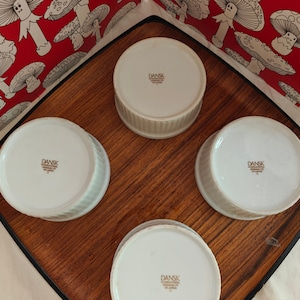 May include: Four white ceramic ramekins with fluted sides, each featuring a brown stamp on the bottom that reads DANSK INTERNATIONAL DESIGNS LTD. SRI LANKA. The ramekins are arranged in a square formation on a wooden tray with a black border.