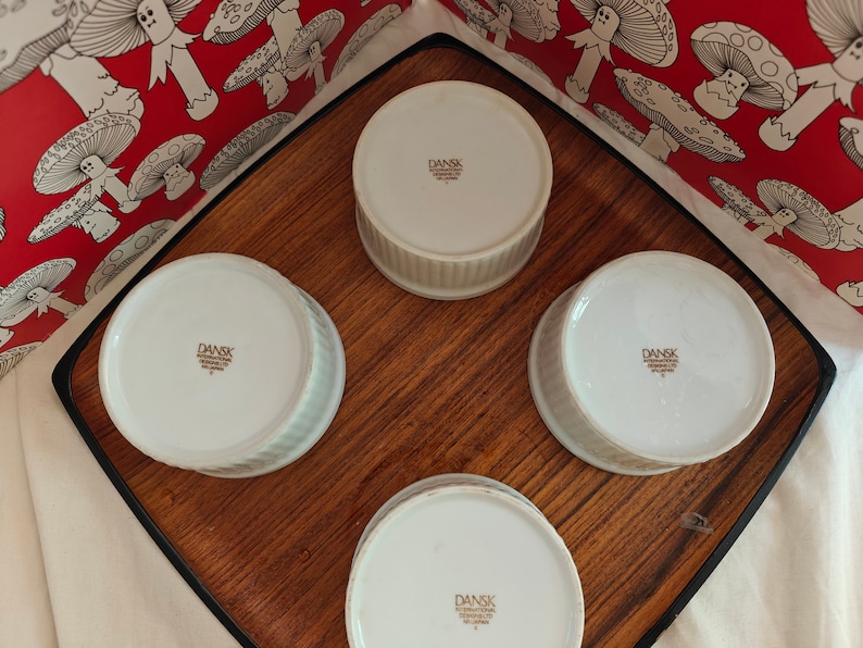 May include: Four white ceramic ramekins with fluted sides, each featuring a brown stamp on the bottom that reads DANSK INTERNATIONAL DESIGNS LTD. SRI LANKA. The ramekins are arranged in a square formation on a wooden tray with a black border.