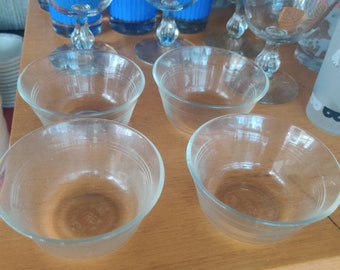 May include: A collection of vintage glassware, including clear glass bowls and stemmed glasses. Some glasses have a gold rim. Blue and red patterned glasses are visible in the background. The items are arranged on a wooden surface.