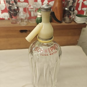 May include: A vintage clear glass soda siphon bottle with vertical fluting and a cream-colored plastic top mechanism. The front of the bottle features the red text SCHWEPPES SODA WATER. It includes a silver-toned metal cap and a black lever.