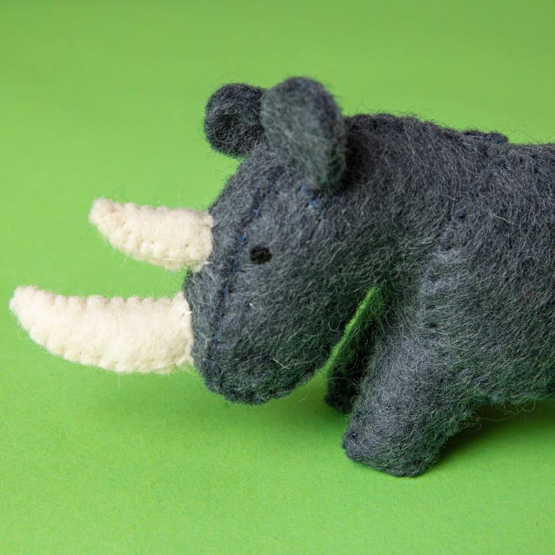 May include: A handcrafted felt rhino toy in grey hues with cream-coloured horns. The rhino features small black eyes and is set against a vibrant green backdrop. This soft sculpture is a delightful addition to a child's toy collection.