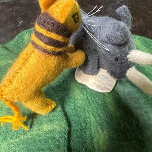 May include: Handmade felted animal figurines. A yellow tiger with a brown striped collar and a grey elephant with white tusks are positioned on a green felt surface. The tiger is facing the elephant.