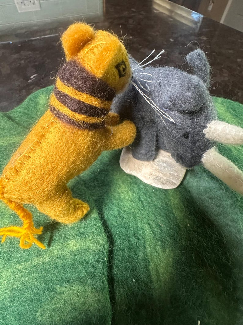 May include: Handmade felted animal figurines. A yellow tiger with a brown striped collar and a grey elephant with white tusks are positioned on a green felt surface. The tiger is facing the elephant.
