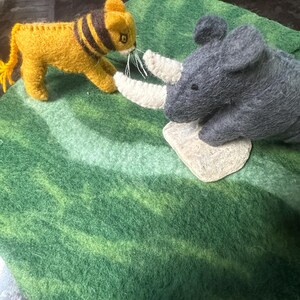 May include: A felt toy scene showing a yellow lion with black stripes and a grey rhinoceros with white horns. They are positioned on a green felt surface with lighter green stripes.