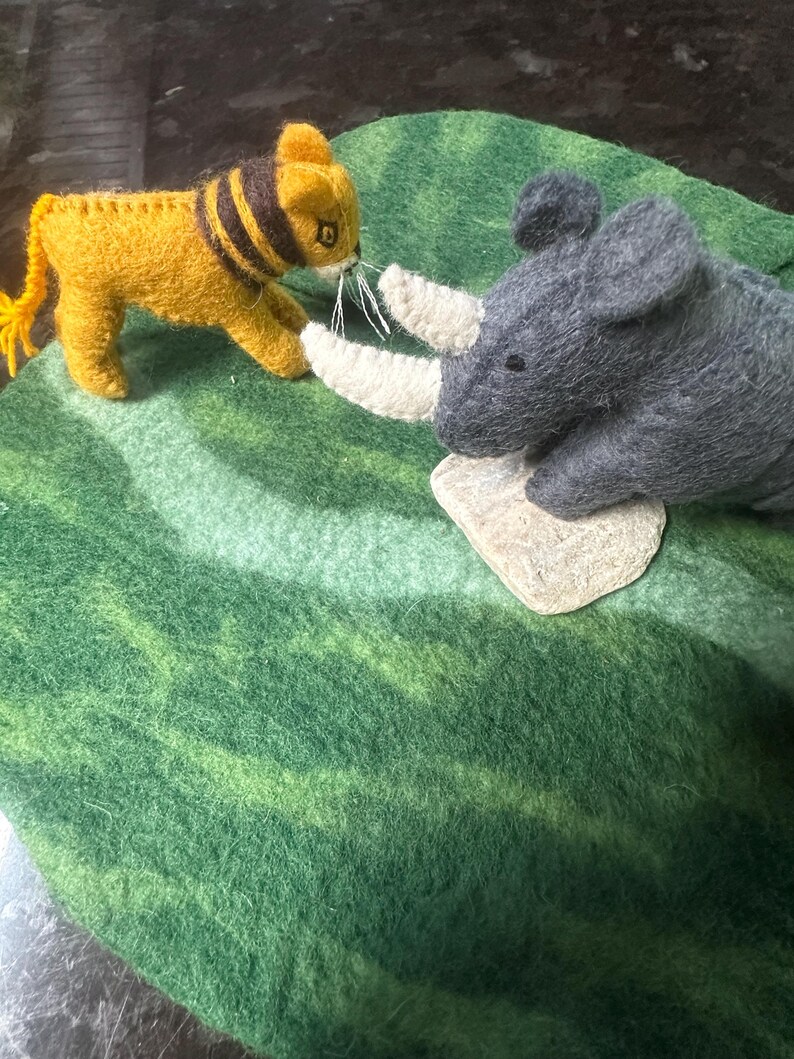 May include: A felt toy scene showing a yellow lion with black stripes and a grey rhinoceros with white horns. They are positioned on a green felt surface with lighter green stripes.
