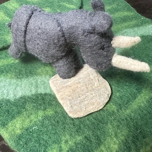 May include: A handcrafted grey felt rhinoceros toy with white horns sits on a small, pale stone. The rhino is placed on a round, green felt mat with a mottled pattern, creating a naturalistic scene.