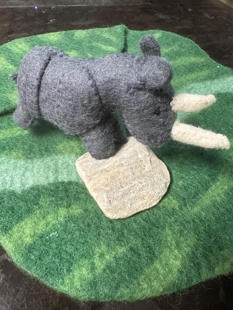 May include: A handcrafted grey felt rhinoceros toy with white horns sits on a small, pale stone. The rhino is placed on a round, green felt mat with a mottled pattern, creating a naturalistic scene.