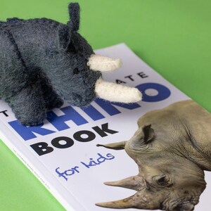 May include: A dark grey felt rhino toy with cream horns sits on a book titled "The Ultimate Rhino Book for Kids." The book cover has a rhino illustration and blue text on a white background. The background is green.