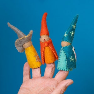 Op de afbeelding: Drie vingerpoppen van vilt, een met een grijze hoed, een met een rood-oranje hoed en een met een blauwe hoed met witte sterren. De poppen zitten op een hand.