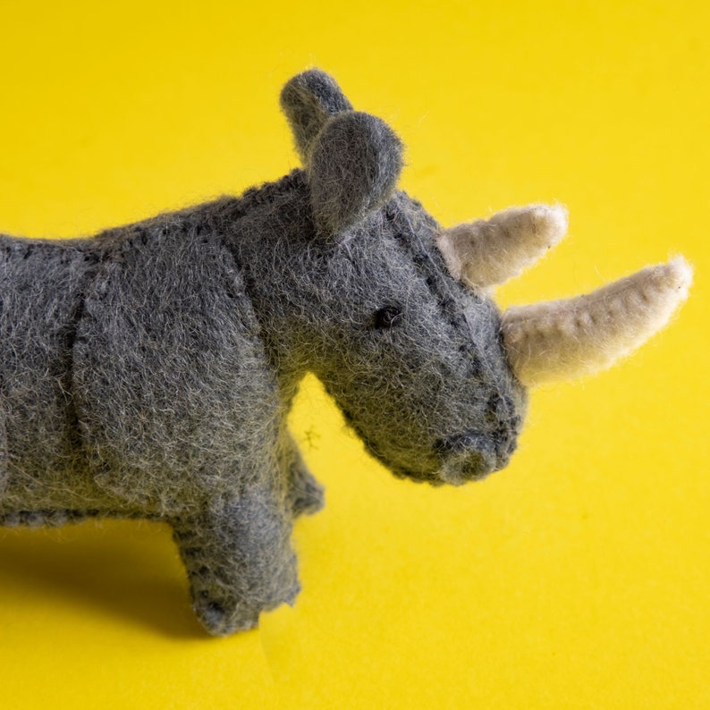 May include: A handcrafted grey felt rhinoceros toy with two cream-coloured horns and small black eyes. The toy is set against a bright yellow background, showcasing its detailed stitching and soft texture. A charming, handcrafted animal figure.