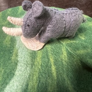 May include: A handcrafted grey felt rhinoceros toy with two cream horns, set on a green felt base. The rhino features small black eyes and a small tail. The felt has a textured finish, giving the toy a soft appearance.