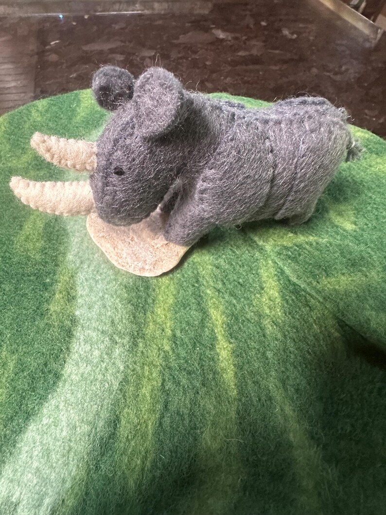May include: A handcrafted grey felt rhinoceros toy with two cream horns, set on a green felt base. The rhino features small black eyes and a small tail. The felt has a textured finish, giving the toy a soft appearance.