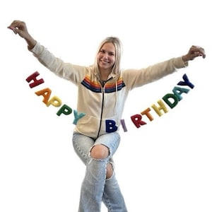 May include: A woman holds up a rainbow felt banner that reads "Happy Birthday".