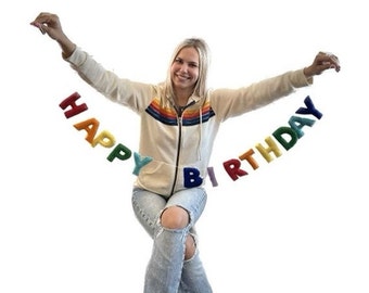 May include: A woman holds up a rainbow felt banner that reads "Happy Birthday".