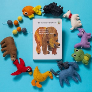 May include: A children's book titled "Brown Bear, Brown Bear, What Do You See?" is surrounded by felt animal ornaments, including a bear, fish, dog, and frog. The book cover features a brown bear illustration. The ornaments are in various colors, arranged on a blue background.