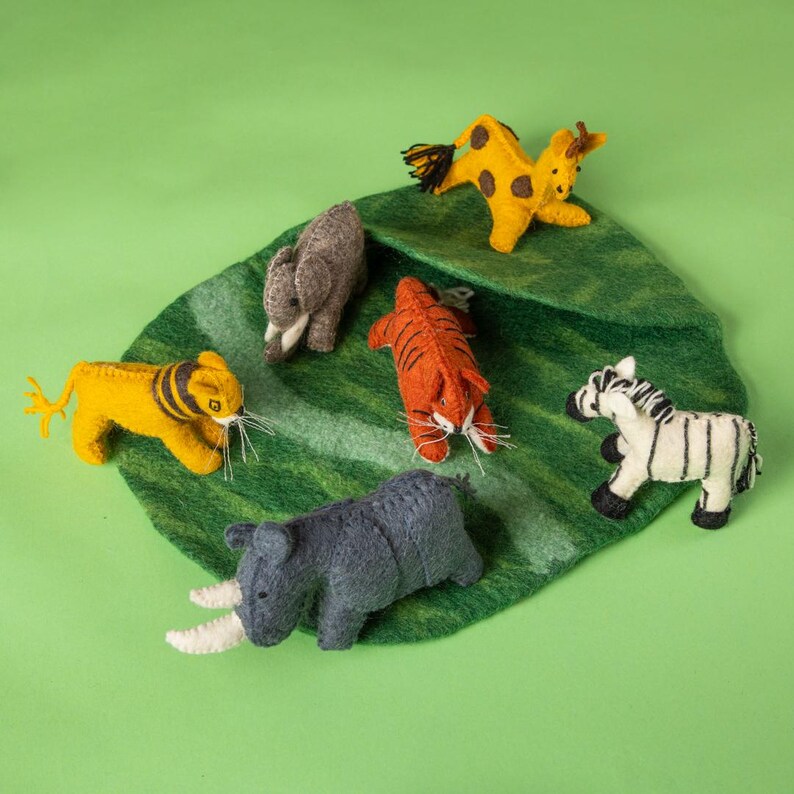 May include: A selection of handmade felt animal figurines, including a yellow lion, orange tiger, grey rhino, zebra, giraffe, and elephant, arranged on a green felt leaf. The animals have detailed features and are set against a solid green background.