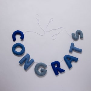 K&ouml;nnte beinhalten: Ein blaues Filzbanner mit dem Wort "CONGRATS" auf einem wei&szlig;en Faden.