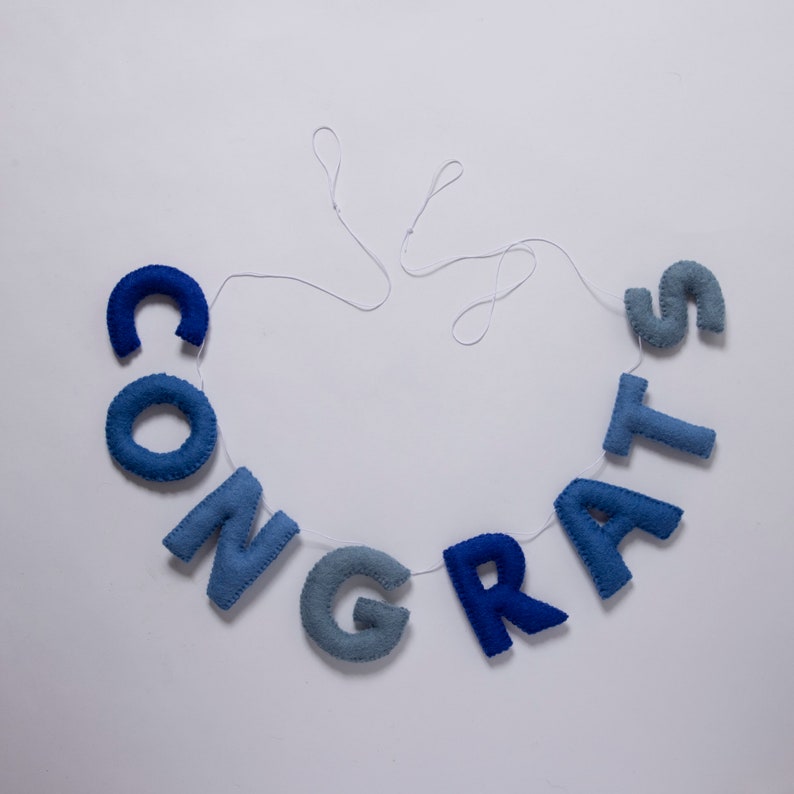 K&ouml;nnte beinhalten: Ein blaues Filzbanner mit dem Wort "CONGRATS" auf einem wei&szlig;en Faden.