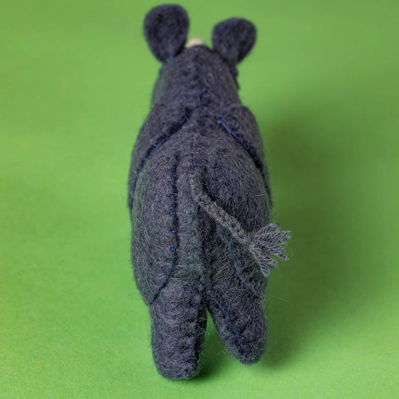 May include: A handmade, dark grey felt rhino toy, seen from the rear, with visible stitching and a small tail. The rhino has small ears and is set against a solid green background. This soft sculpture is a unique children's toy.