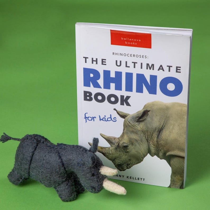 May include: A dark grey felt rhino toy and a book titled "The Ultimate Rhino Book for kids." The book cover shows a rhino illustration. The toy has white tusks and a small tail. The background is green.