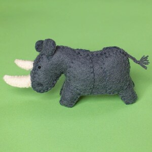 May include: A handcrafted, grey felt rhinoceros toy with two white horns, small black eyes, and a braided tail. The toy is set against a solid green background. This soft sculpture is a charming addition to a child's toy collection.