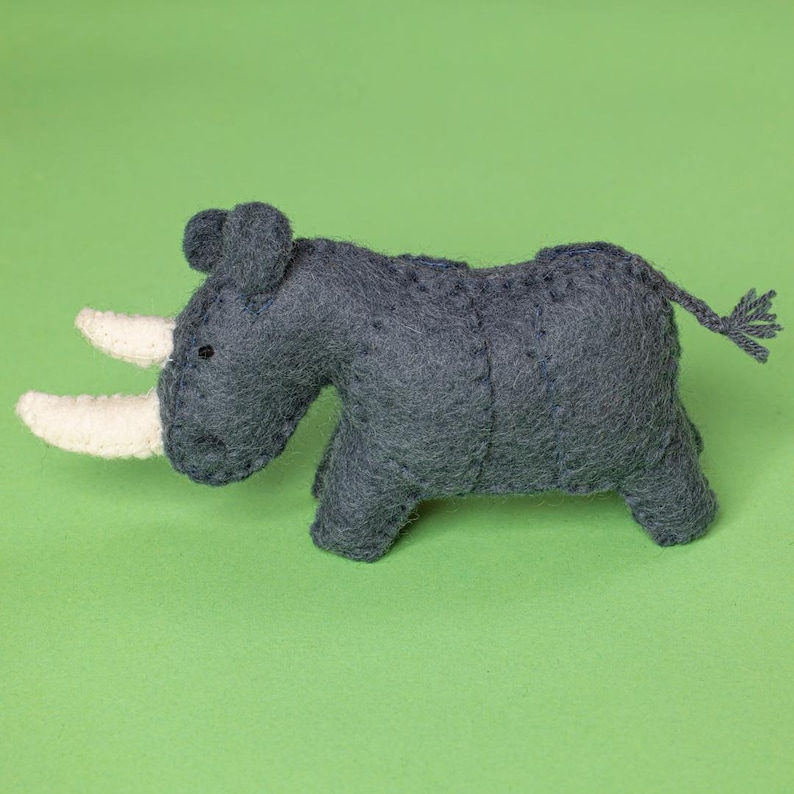 May include: A handcrafted, grey felt rhinoceros toy with two white horns, small black eyes, and a braided tail. The toy is set against a solid green background. This soft sculpture is a charming addition to a child's toy collection.