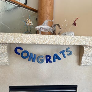 K&ouml;nnte beinhalten: Ein blaues Filzbanner mit dem Wort "CONGRATS" h&auml;ngt &uuml;ber einem Kamin. Das Banner besteht aus einzelnen Buchstaben, die jeweils einen anderen Blauton haben.
