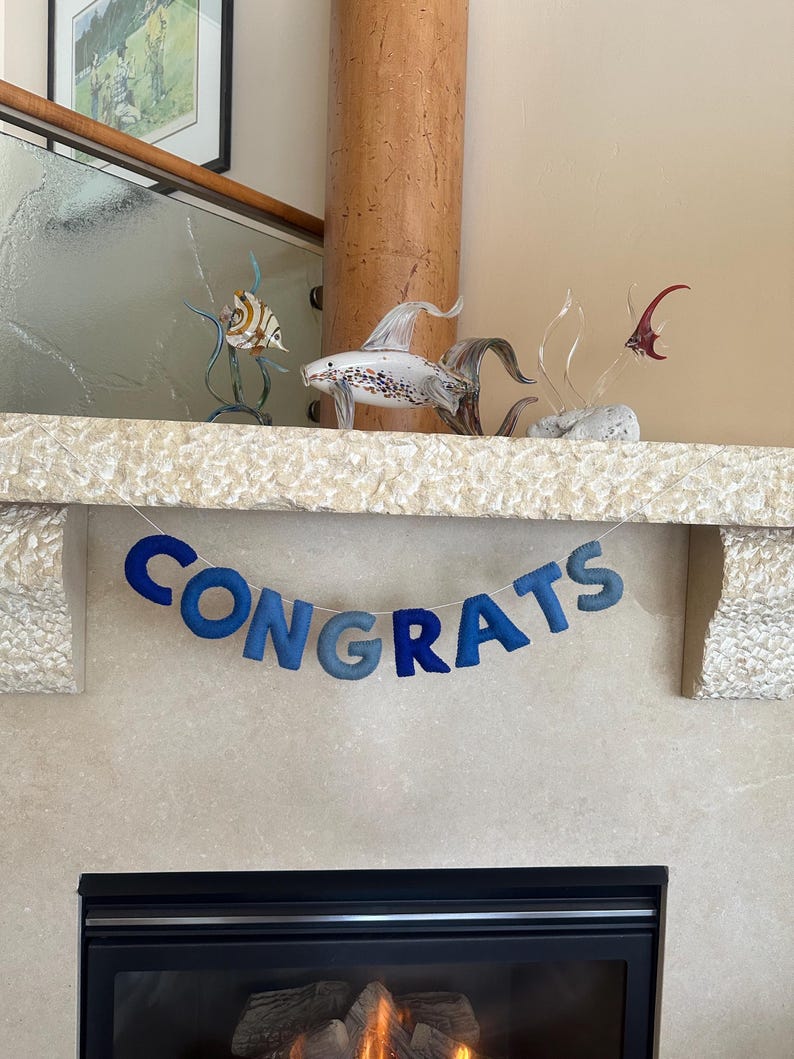 K&ouml;nnte beinhalten: Ein blaues Filzbanner mit dem Wort "CONGRATS" h&auml;ngt &uuml;ber einem Kamin. Das Banner besteht aus einzelnen Buchstaben, die jeweils einen anderen Blauton haben.