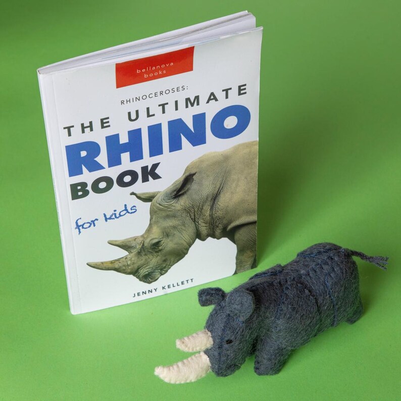 May include: A children's book titled "The Ultimate Rhino Book" with a rhino illustration, next to a dark grey stuffed rhino toy with white horns, set against a green background.