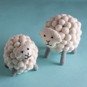 May include: Two white felt sheep with black eyes and noses. The sheep have a fluffy texture and are standing on a light blue background.