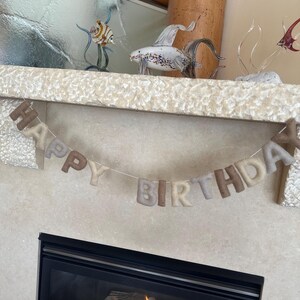 May include: A felt banner with the words "Happy Birthday" in beige and brown letters hanging above a fireplace with a fire burning inside.
