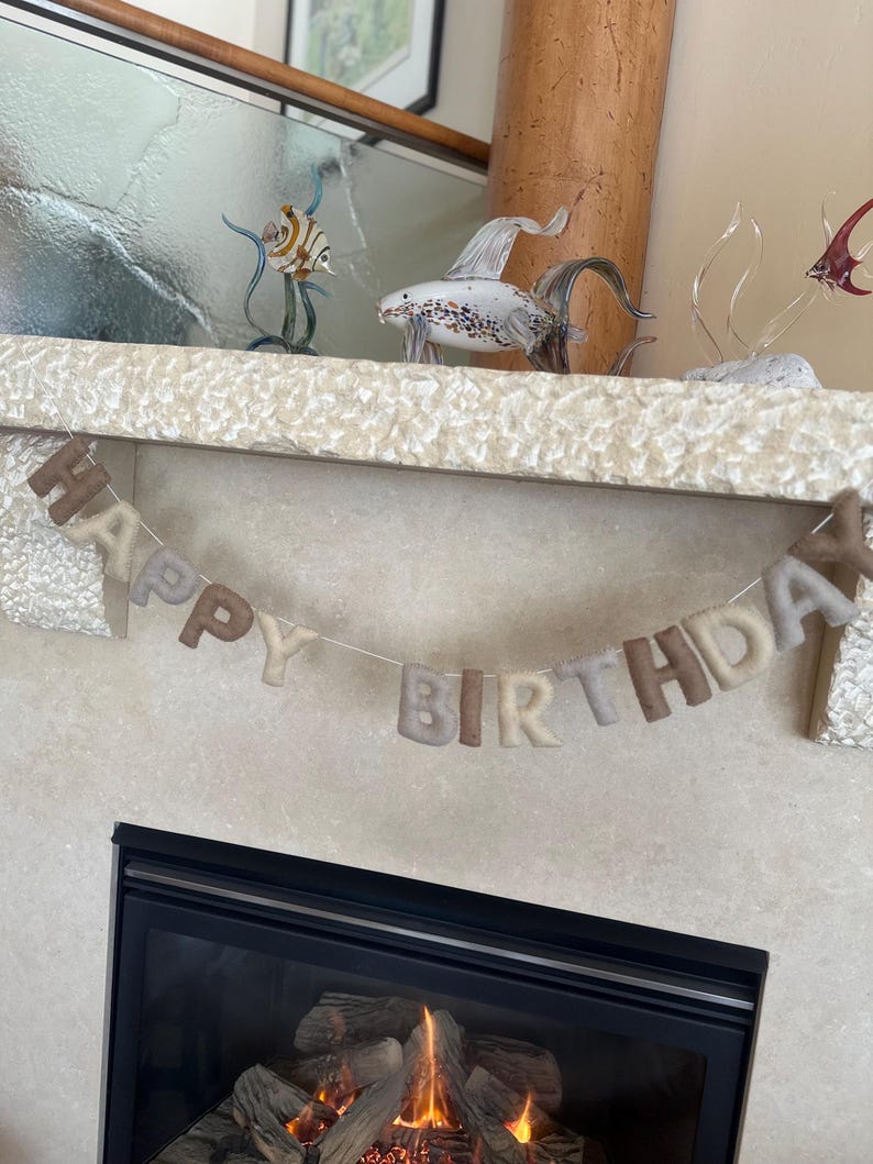 May include: A felt banner with the words "Happy Birthday" in beige and brown letters hanging above a fireplace with a fire burning inside.
