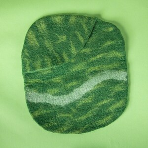 May include: A handmade, oval-shaped felt item in shades of green and yellow, with a wavy, light grey stripe across the centre. The felt has a textured appearance, and the item is set against a solid green background.