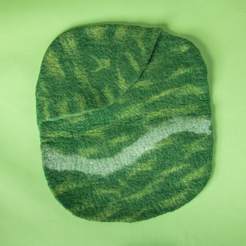 May include: A handmade, oval-shaped felt item in shades of green and yellow, with a wavy, light grey stripe across the centre. The felt has a textured appearance, and the item is set against a solid green background.