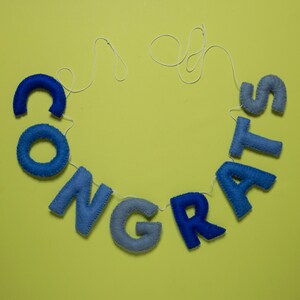 K&ouml;nnte beinhalten: Ein blaues Filzbanner mit dem Wort "CONGRATS" auf einem wei&szlig;en Faden vor gelbem Hintergrund.