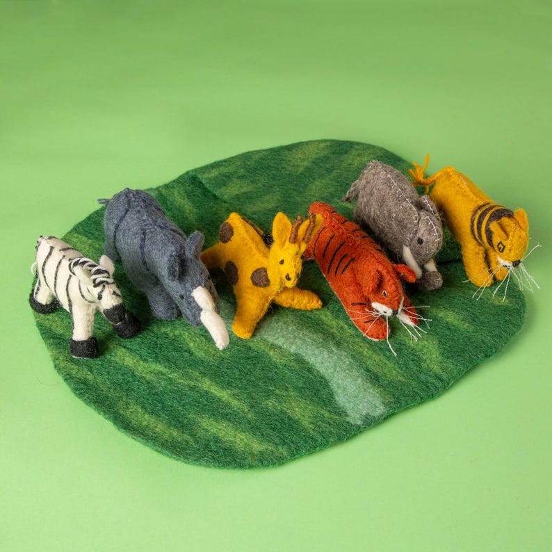 May include: A selection of handmade felt animal toys, including a zebra, rhino, giraffe, tiger, and rabbit, displayed on a green leaf-shaped mat. The animals are in various colours, such as white, black, yellow, orange, and grey.