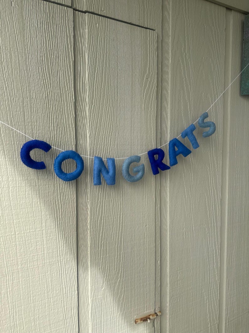 K&ouml;nnte beinhalten: Ein blaues Filzbanner mit dem Wort "CONGRATS" in einzelnen Buchstaben geschrieben. Die Buchstaben sind mit wei&szlig;em Garn zusammengef&uuml;gt und h&auml;ngen an einer hellgrauen Holzwand.