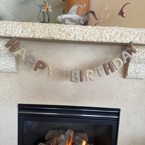 May include: A felt banner with the words "Happy Birthday" in brown and beige letters, hanging above a fireplace with a fire burning inside.