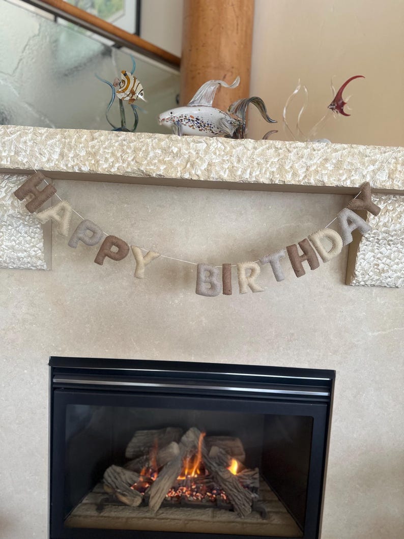 May include: A felt banner with the words "Happy Birthday" in brown and beige letters, hanging above a fireplace with a fire burning inside.