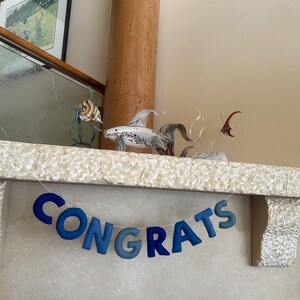 K&ouml;nnte beinhalten: Ein blaues Filzbanner mit dem Wort "CONGRATS" h&auml;ngt &uuml;ber einem Kamin. Das Banner ist mit wei&szlig;em Garn befestigt. Zwei gl&auml;serne Fischfiguren stehen auf dem Kaminsims.