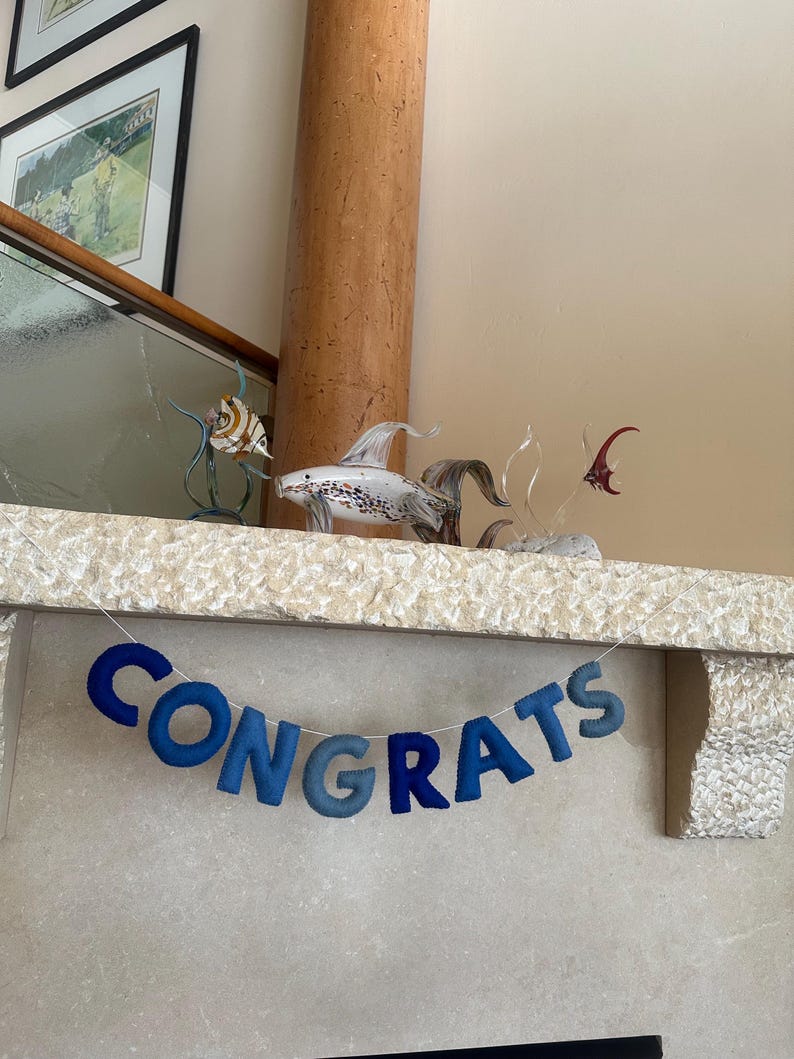 K&ouml;nnte beinhalten: Ein blaues Filzbanner mit dem Wort "CONGRATS" h&auml;ngt &uuml;ber einem Kamin. Das Banner ist mit wei&szlig;em Garn befestigt. Zwei gl&auml;serne Fischfiguren stehen auf dem Kaminsims.
