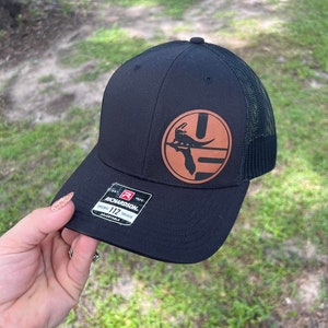 May include: Black and brown baseball cap with a brown leather patch featuring a black alligator silhouette and the state of Florida. The patch is on the front of the cap. The cap is a trucker style with a mesh back.