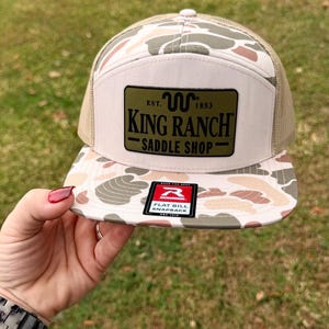 Könnte beinhalten: Eine Flat-Bill-Snapback-Kappe mit Tarnmuster auf dem Schirm und einem beigefarbenen Mesh-Rücken. Die Vorderseite ist beige mit einem grünen rechteckigen Aufnäher mit dem schwarzen Text "KING RANCH SADDLE SHOP".