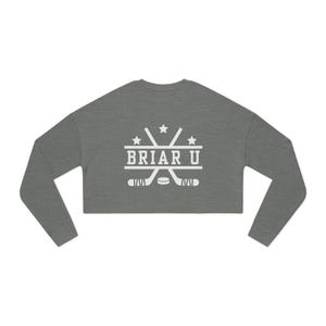 Puede incluir: Una sudadera corta gris jaspeado con mangas largas. La parte delantera presenta el texto blanco "BRIAR U" con palos de hockey cruzados y estrellas. La sudadera es de estilo atlético informal.