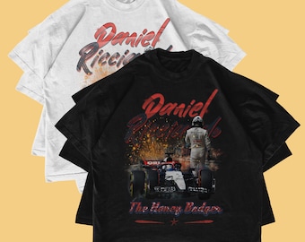 May include: Two t-shirts, one white and one black, featuring a design with the name "Daniel Ricciardo" in red script. The black shirt has an image of a race car and the text "The Honey Badger."