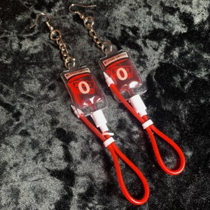 May include: A pair of novelty earrings featuring miniature blood bags. Each earring has a clear plastic blood bag with red liquid, the text "BLOOD GROUP O", and a red tube with a loop. The earrings hang from silver-colored chains and hooks.