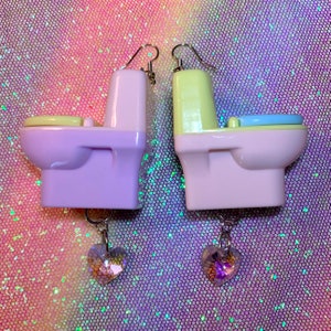 May include: A pair of dangle earrings featuring miniature toilet figurines with a pastel purple and yellow color scheme. Each toilet has a light blue or yellow seat and is accented with a dangling crystal heart charm.