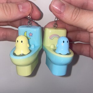 May include: A pair of novelty earrings featuring miniature toilets in pastel colors. One earring has a yellow toilet with a yellow ghost, the other has a blue toilet with a blue ghost. Each toilet has a decorative element.